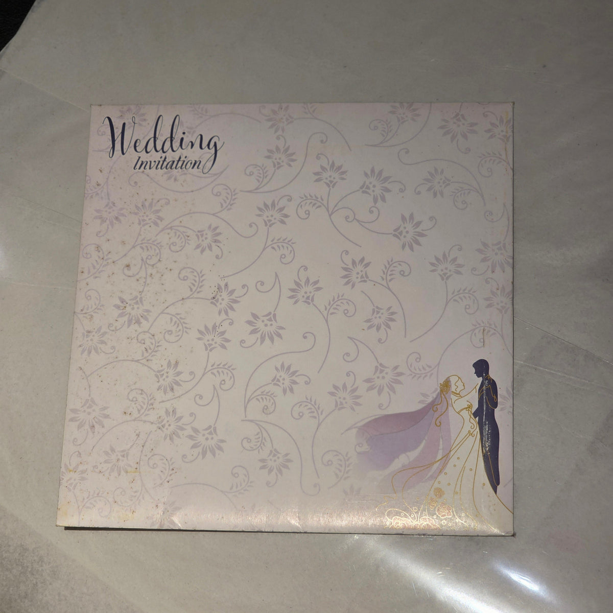 A square wedding invitation card with a pale pink and lavender floral pattern, featuring the text 'Wedding invitation' and an illustration of a bride and groom in the bottom right corner, placed on a light gray surface.