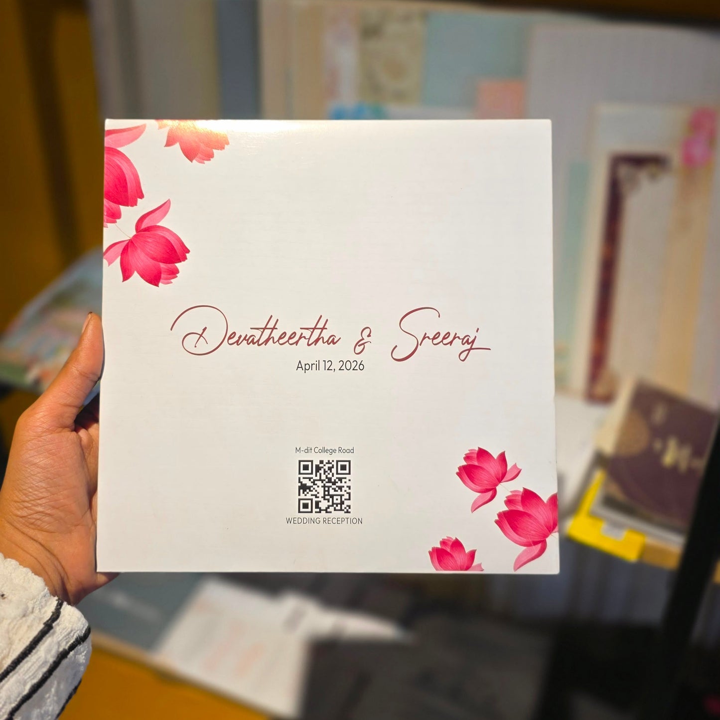 A hand holds a white square wedding program or invitation card featuring pink floral illustrations in the corners and elegant cursive text reading 'Denaturetha & George' with the date 'April 12, 2026'. The background is a softly blurred indoor setting with warm brown tones.