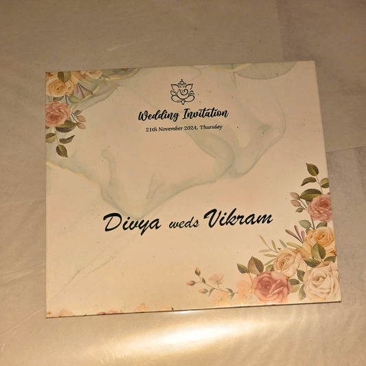 A square wedding invitation card with a cream background, featuring a light blue marble wash in the upper half, floral illustrations in pink, peach, and green in the corners, and the text 'Wedding Invitation' and 'Divya weds Vikram' printed on it.