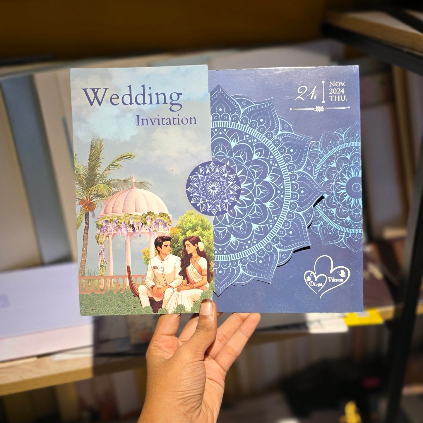 A hand holds two wedding invitation cards in a store setting. The left card is a traditional white card with 'Wedding Invitation' text and a wedding scene, while the right card is a blue mandala-patterned card with a cutout window showing a small white card inside.