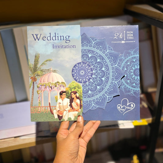 A hand holds two wedding invitation cards in a store setting. The left card is a traditional white card with 'Wedding Invitation' text and a wedding scene, while the right card is a blue mandala-patterned card with a cutout window showing a small white card inside.