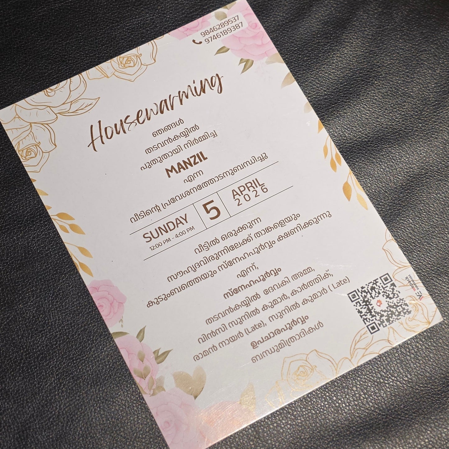 A rectangular printed card with floral pink and gold border design, photographed on a dark textured surface. The card contains event text in English and Thai, including the heading 'Housewarming' and details for a Sunday 5 PM event.