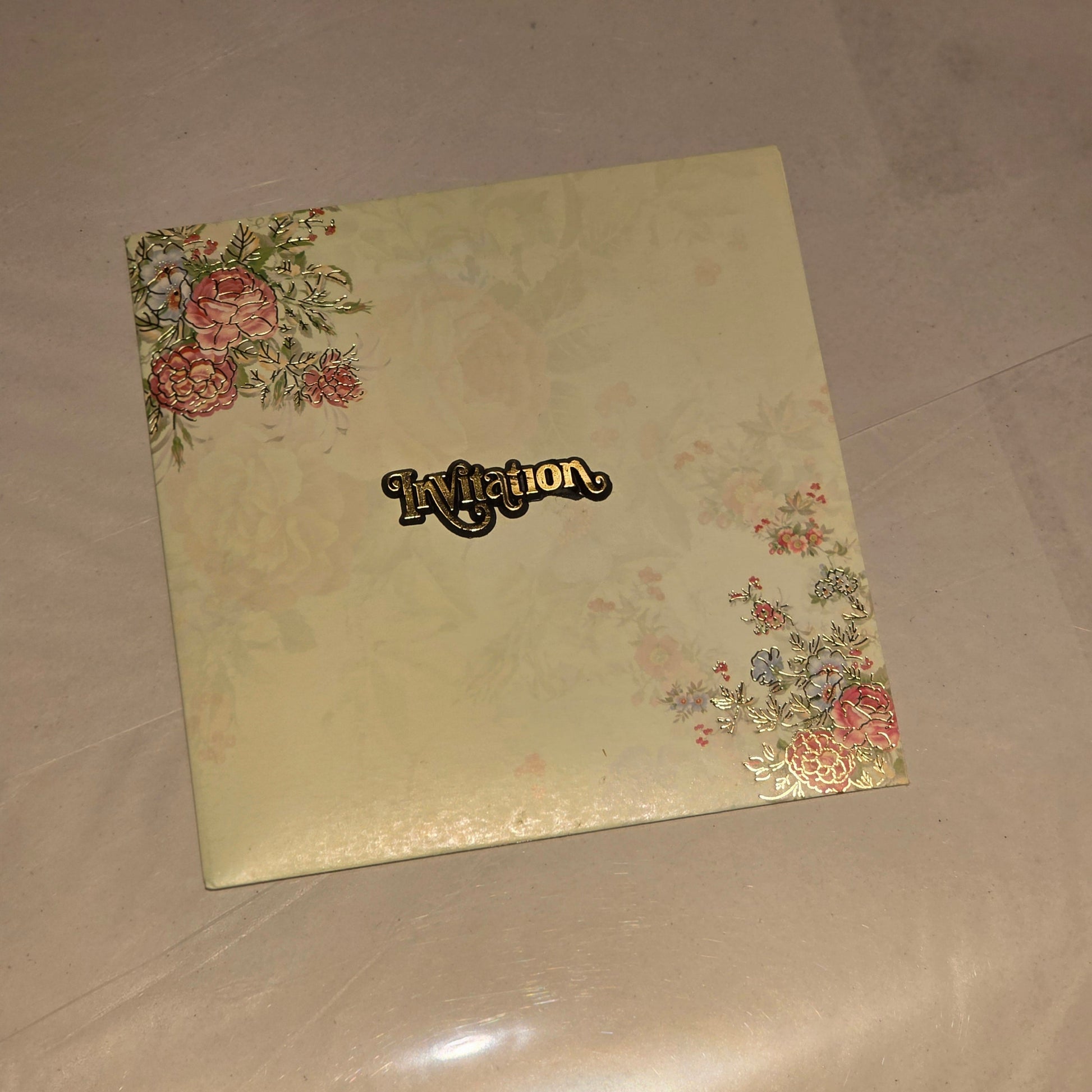 A square cream-colored invitation card featuring a subtle floral pattern in the corners and a black script 'Invitation' label in the center, resting on a light beige surface.
