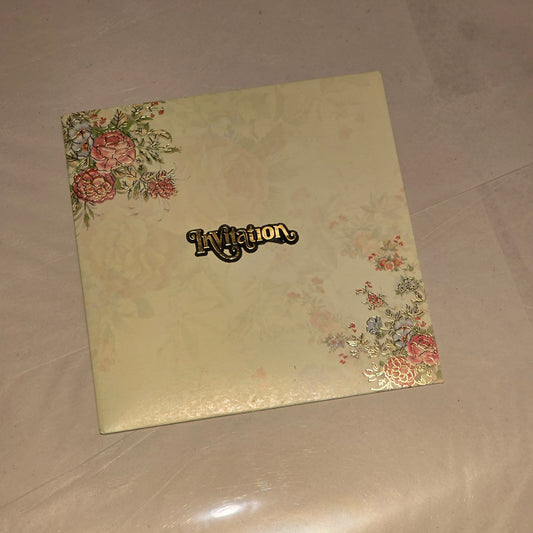 A square cream-colored invitation card featuring a subtle floral pattern in the corners and a black script 'Invitation' label in the center, resting on a light beige surface.