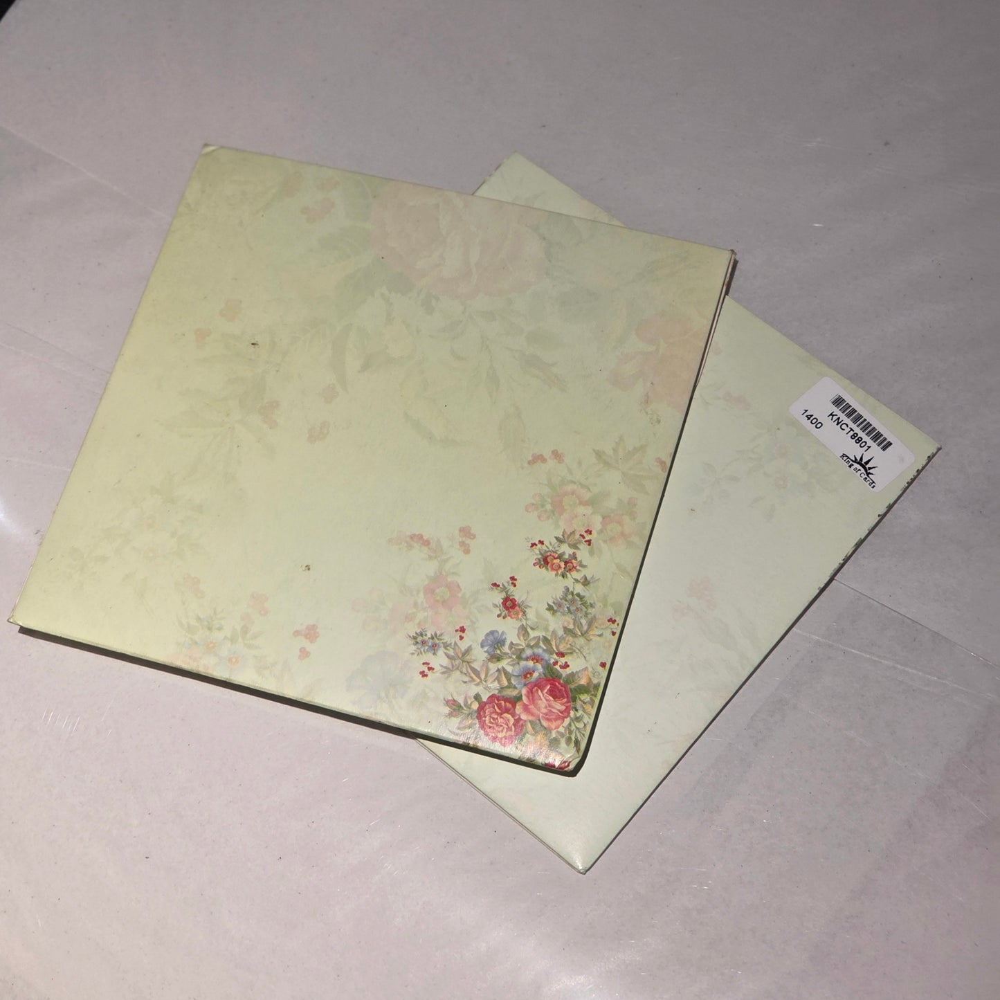 Two square floral paper napkins or small cards with a pale yellow-green background and delicate rose print, stacked on a light fabric surface.