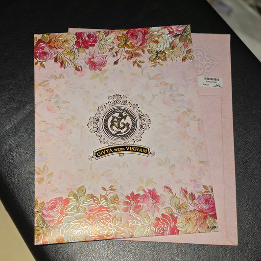 A pink floral invitation card with gold foil accents and a central monogram logo, placed on a black surface next to a matching pink envelope.