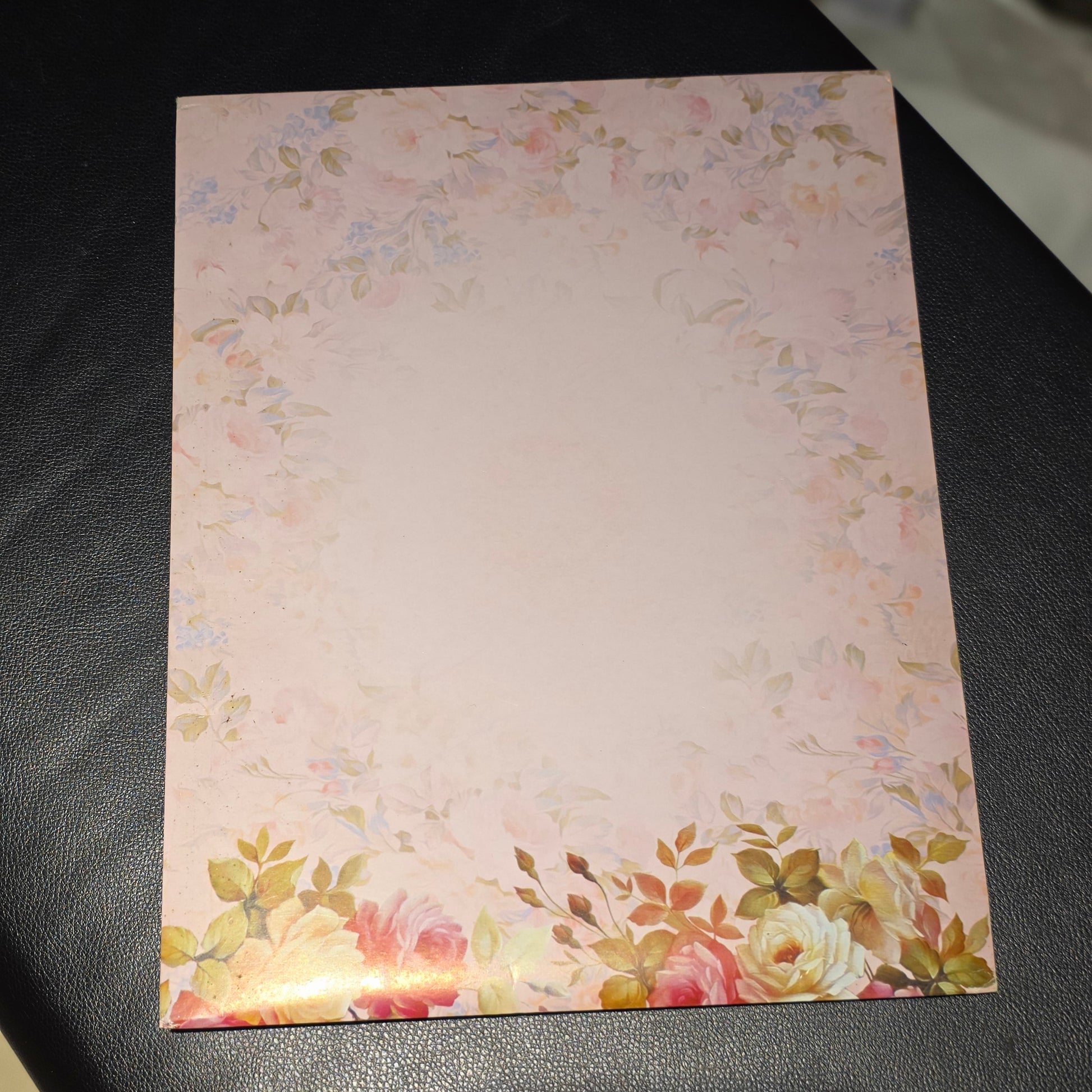 A rectangular floral notepad or card with a pale pink center and a watercolor-style floral border, placed on a dark textured surface.