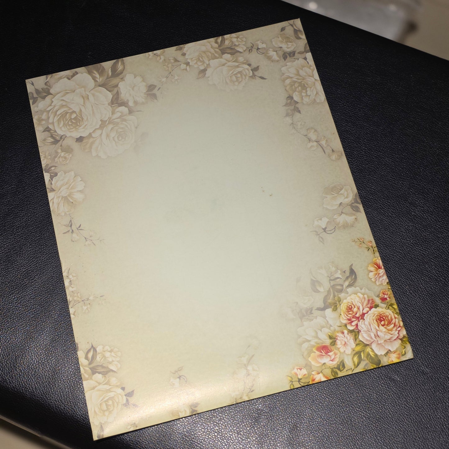 A rectangular card or paper with a vintage floral border design in cream, beige, and soft pink tones, placed on a dark textured surface.