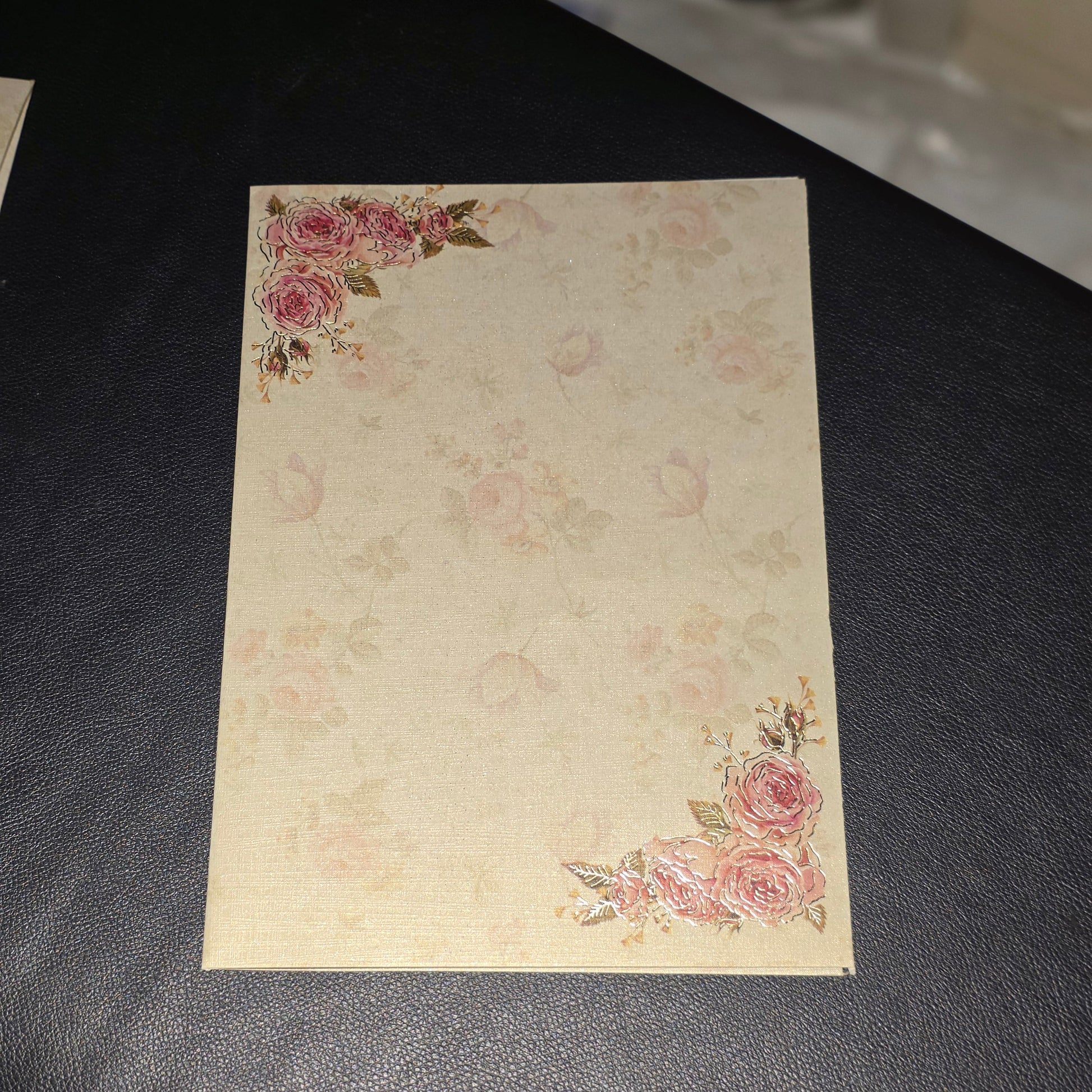 A rectangular cream-colored card or envelope with a vintage floral pattern and pink rose corners, resting on a dark textured surface.