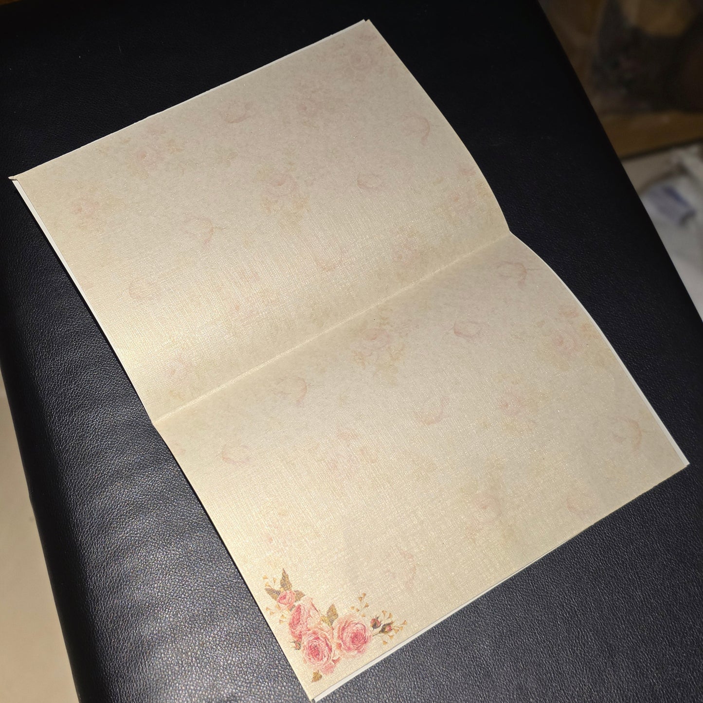 An open cream-colored linen-style notebook or journal with a subtle floral pattern and a small pink rose design in the bottom-left corner, resting on a dark surface.