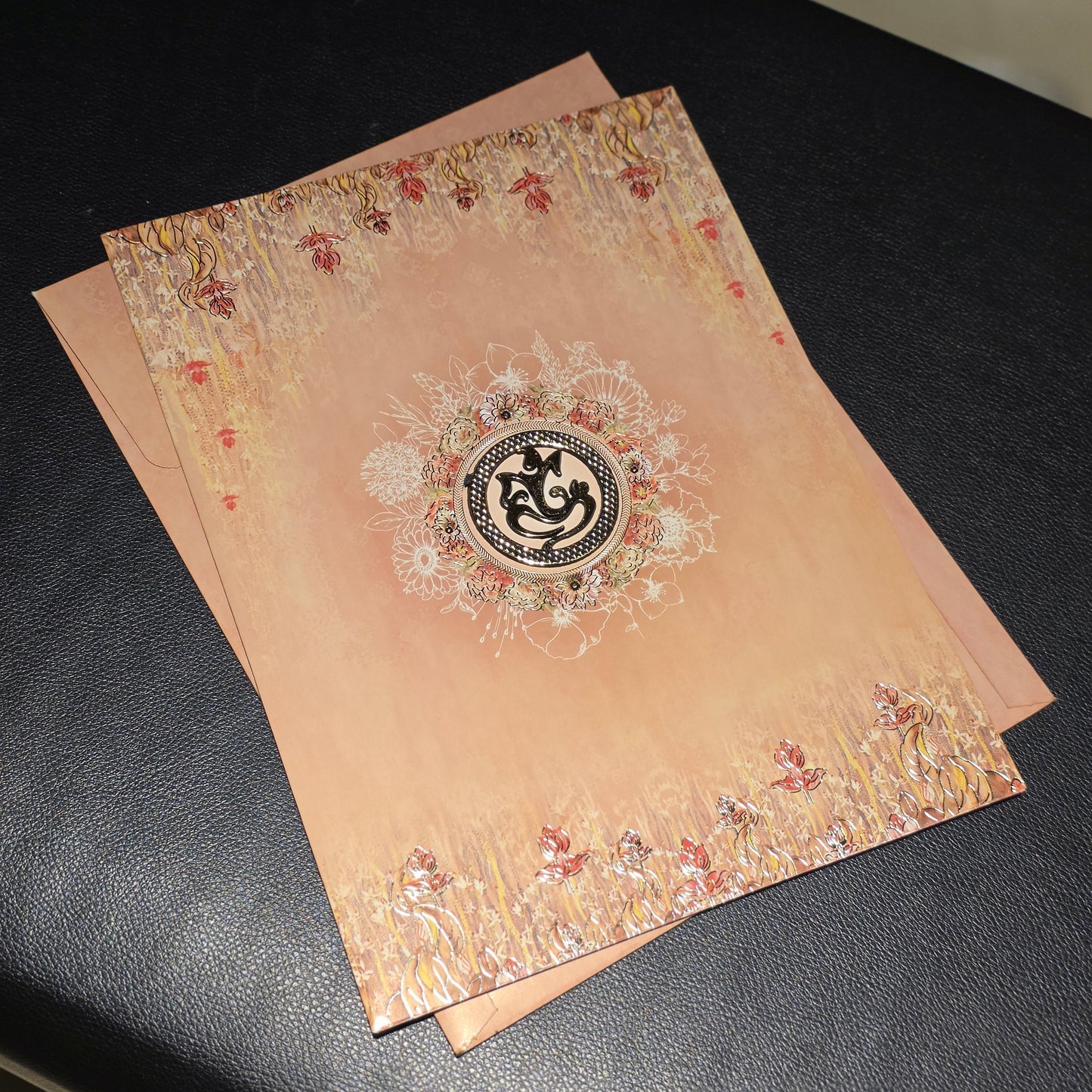 A peach-colored decorative card or invitation with ornate floral borders and a central circular emblem, placed on a black surface.