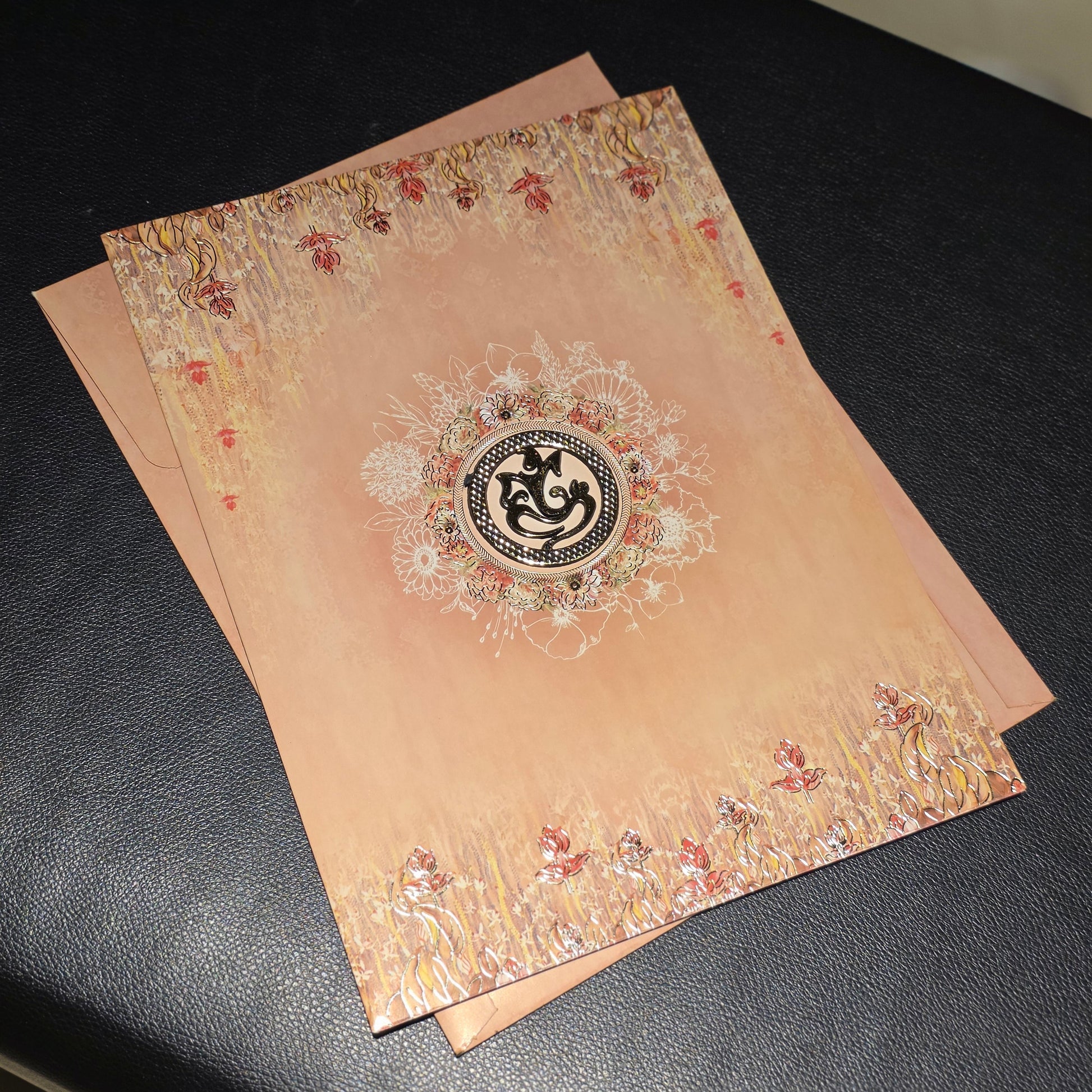 A peach-colored decorative card or invitation with ornate floral borders and a central circular emblem, placed on a black surface.