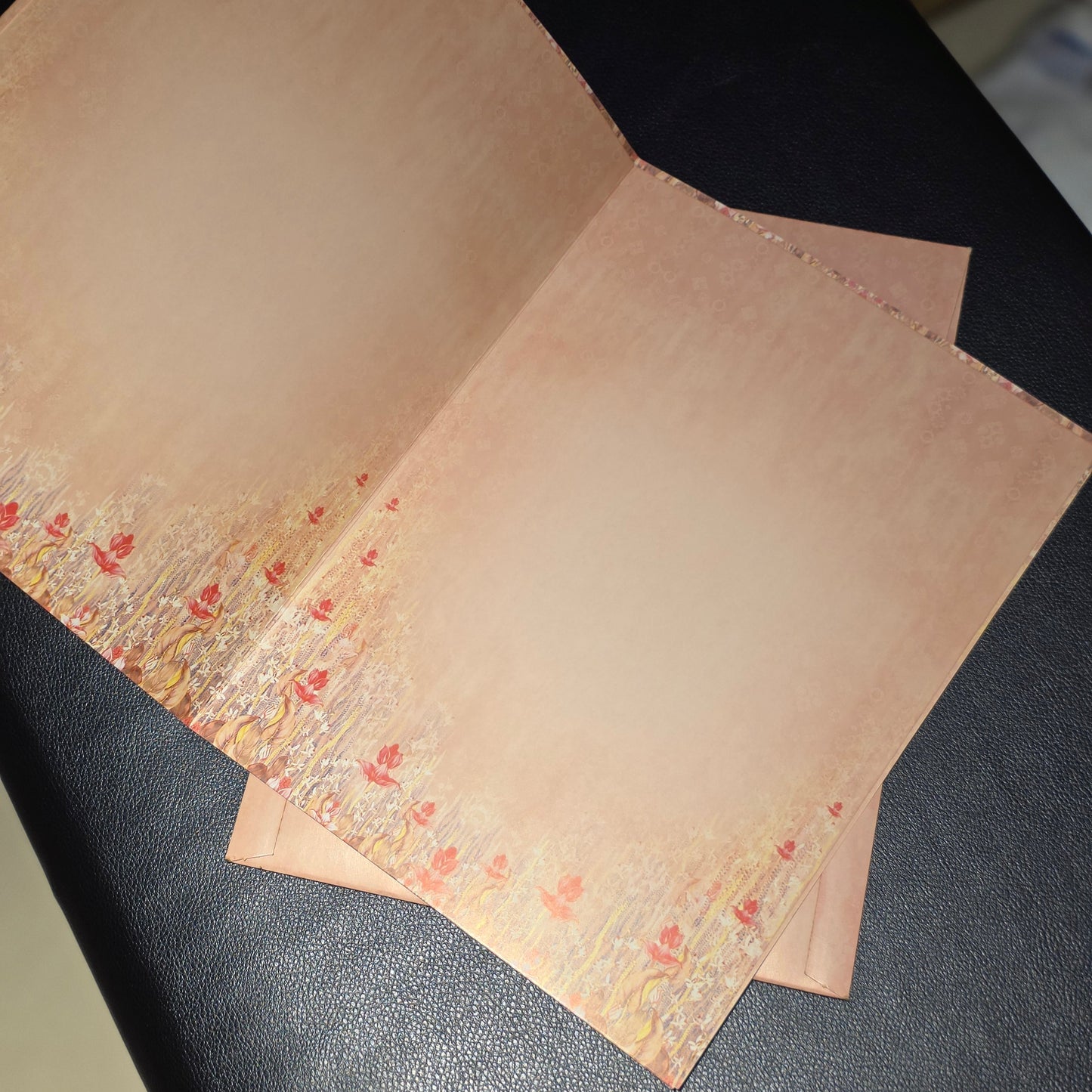 A handmade paper card or invitation with a warm beige background and a delicate red floral border along the bottom edge, displayed on a dark textured surface.