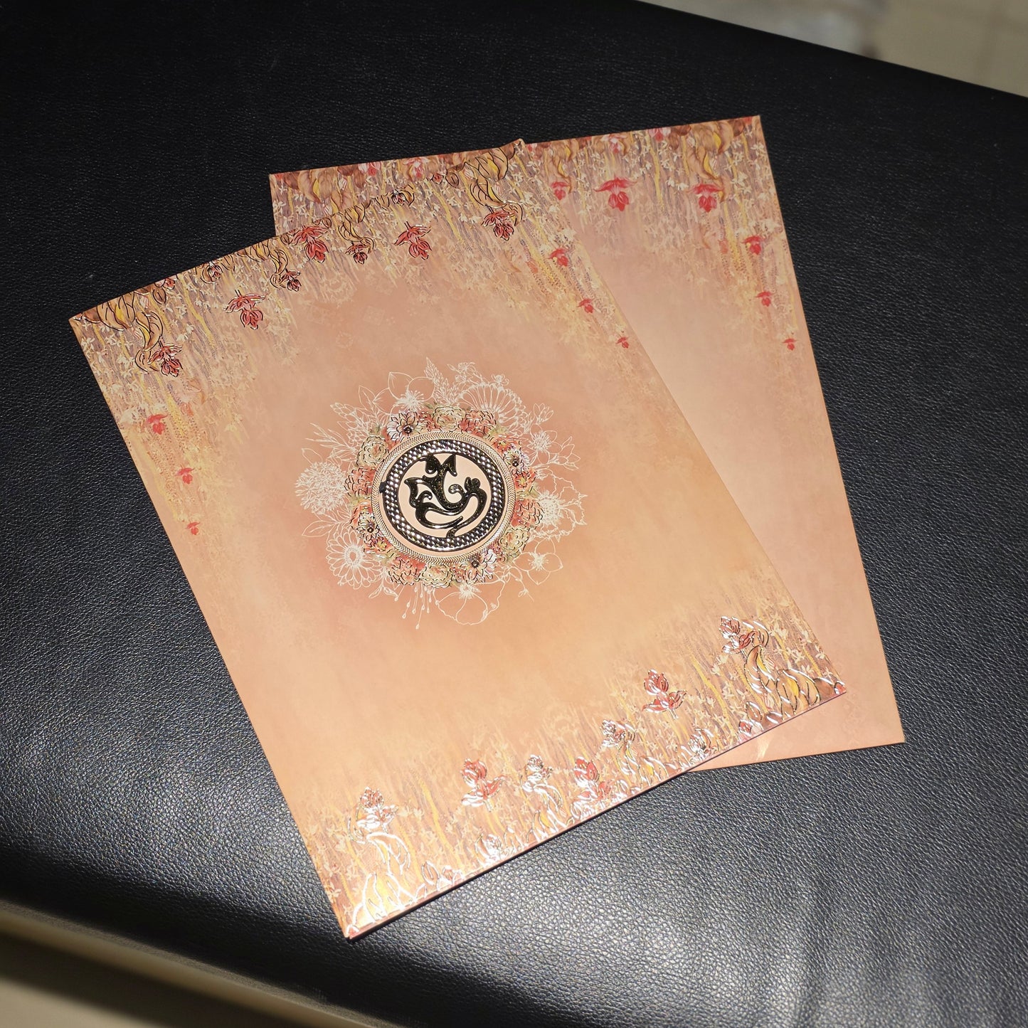 Two decorative peach-colored cards or envelopes with ornate white mandala designs and small red floral accents, placed on a black leather surface.