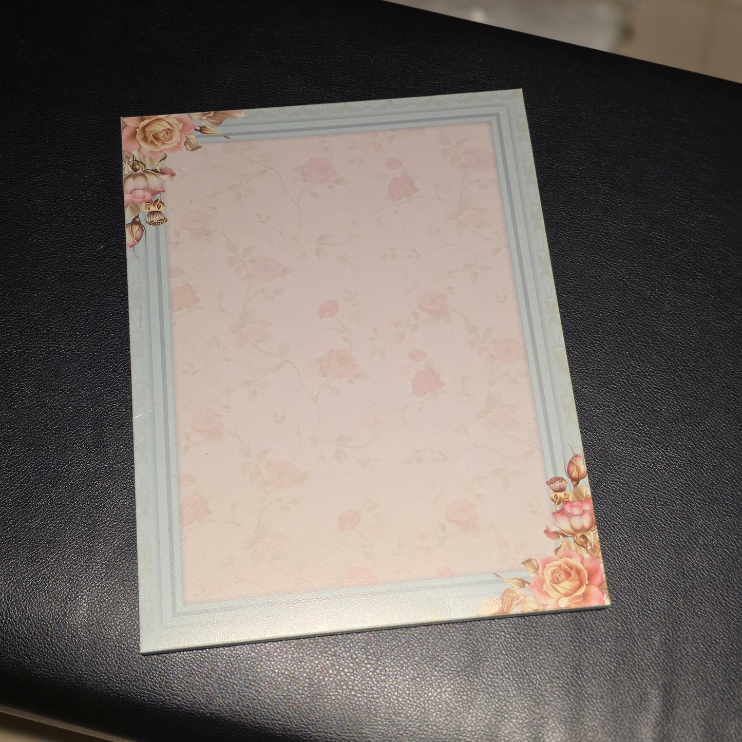A square stationery card or notepad with a pale pink floral pattern, a light blue border, and pink rose corner decorations, placed on a black surface.