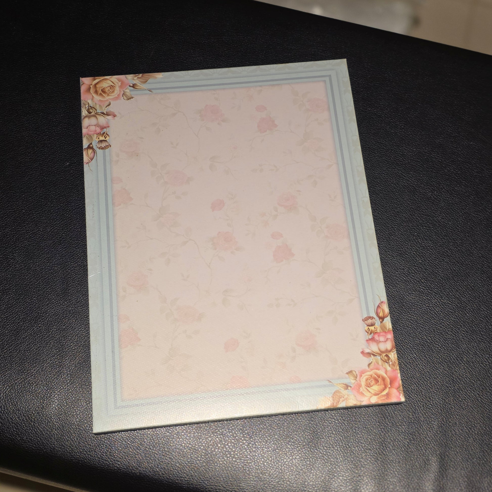 A square stationery card or notepad with a pale pink floral pattern, a light blue border, and pink rose corner decorations, placed on a black surface.