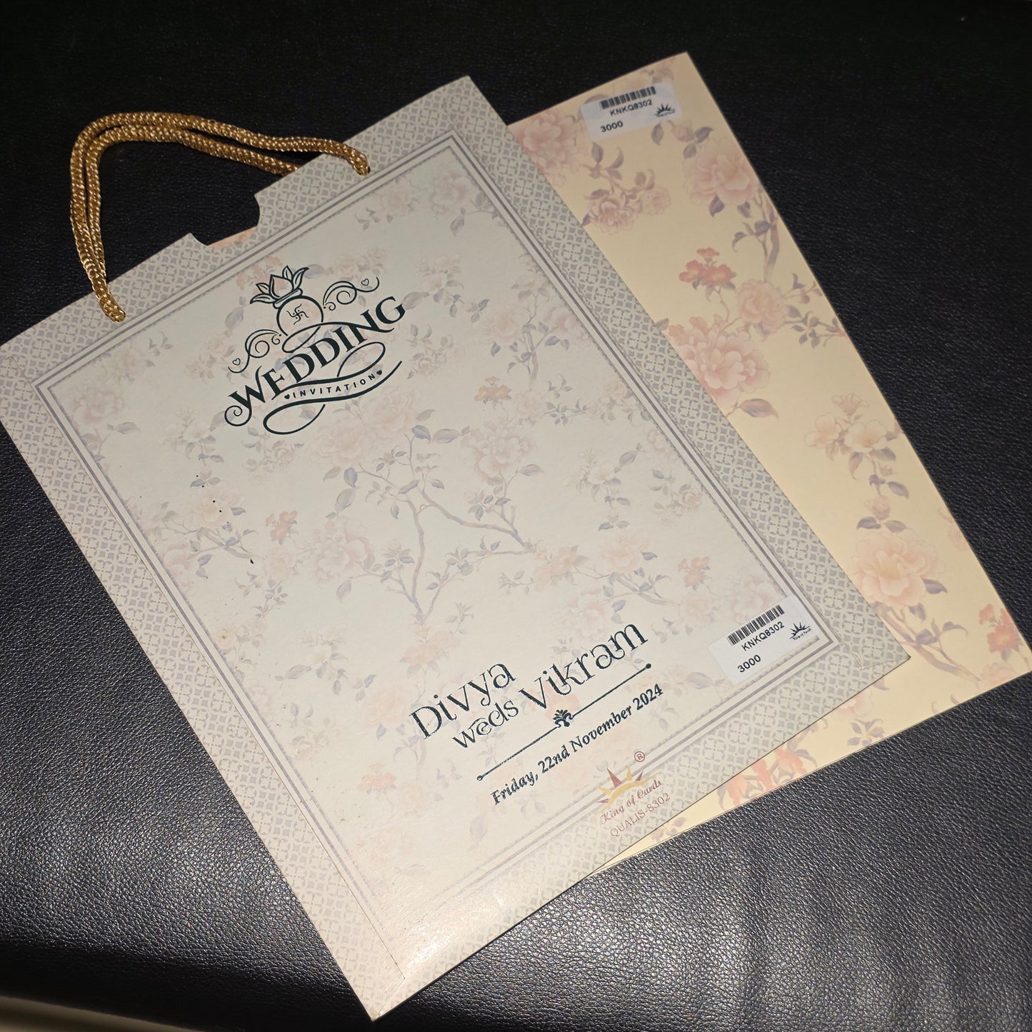 A printed wedding invitation card and matching gift bag with floral designs, placed on a dark textured surface. The bag has gold rope handles and the card reads 'WEDDING' and 'Divya and Vikram'.