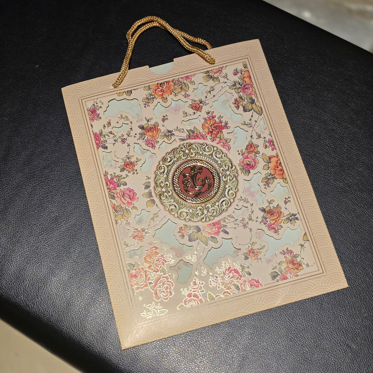 A decorative paper gift bag with gold rope handles, featuring a floral pattern and a central circular emblem, resting on a dark textured surface.