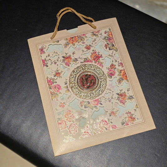 A decorative paper gift bag with gold rope handles, featuring a floral pattern and a central circular emblem, resting on a dark textured surface.