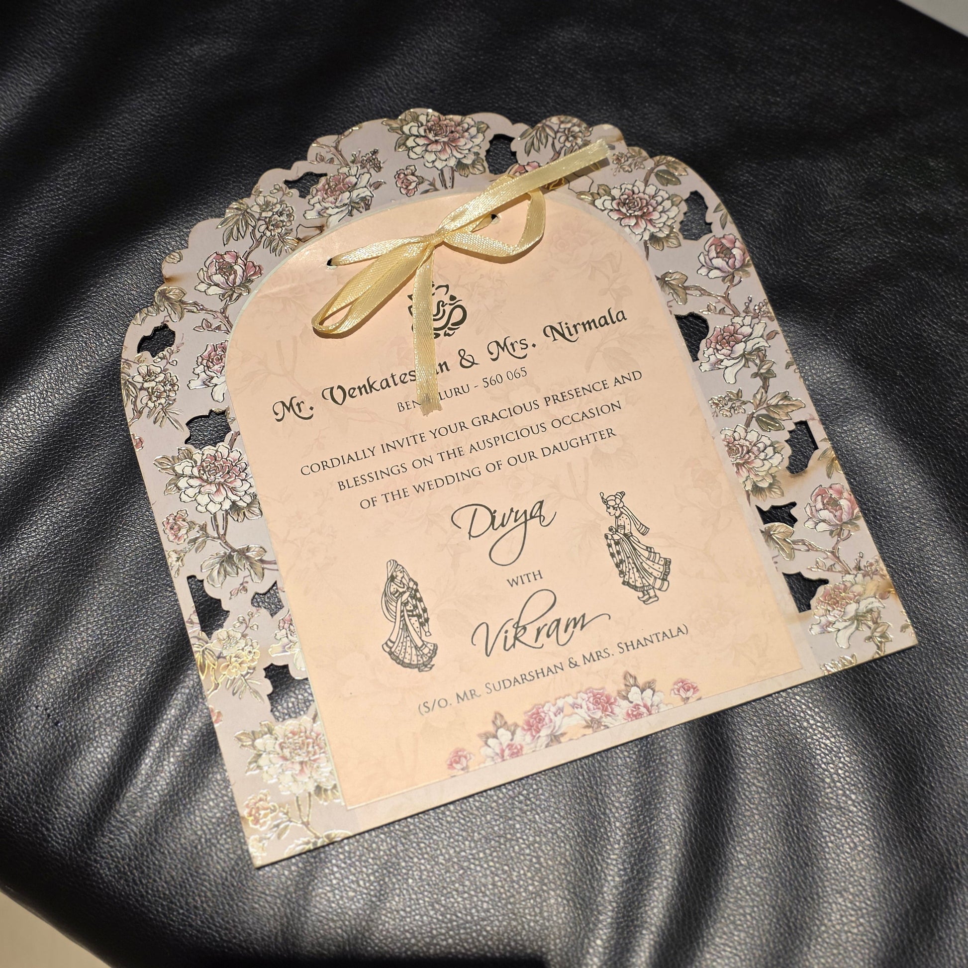 A decorative wedding invitation card with floral lace-like border details and a gold ribbon bow, placed on a black leather surface.
