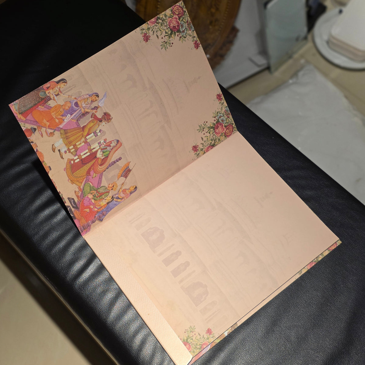 A peach-colored open book or album with traditional illustrated covers, displayed on a black leather surface in a room setting.