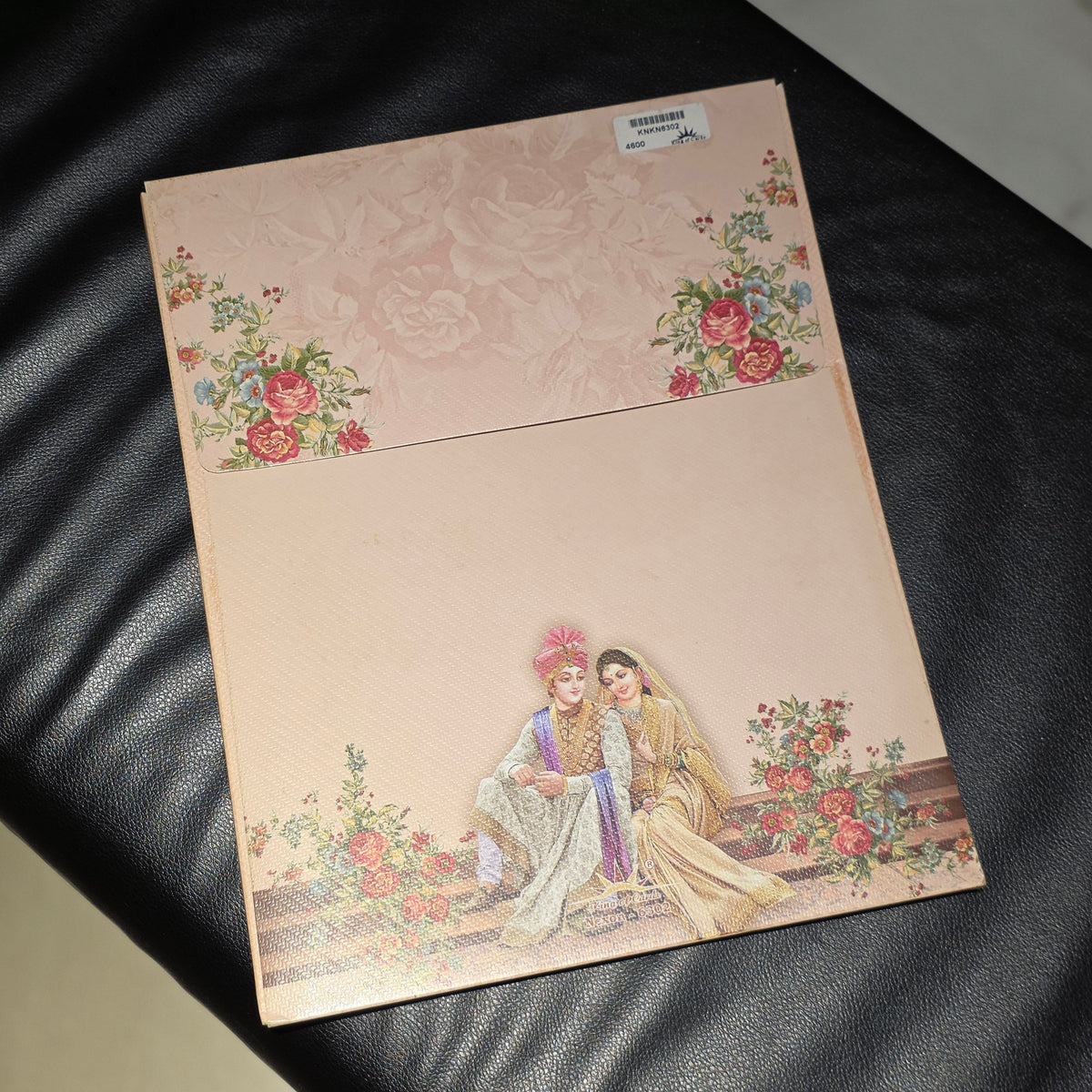 A square wedding invitation card with a light pink background, featuring floral corner designs and an illustration of a bride and groom seated together at the bottom, placed on a black leather surface.