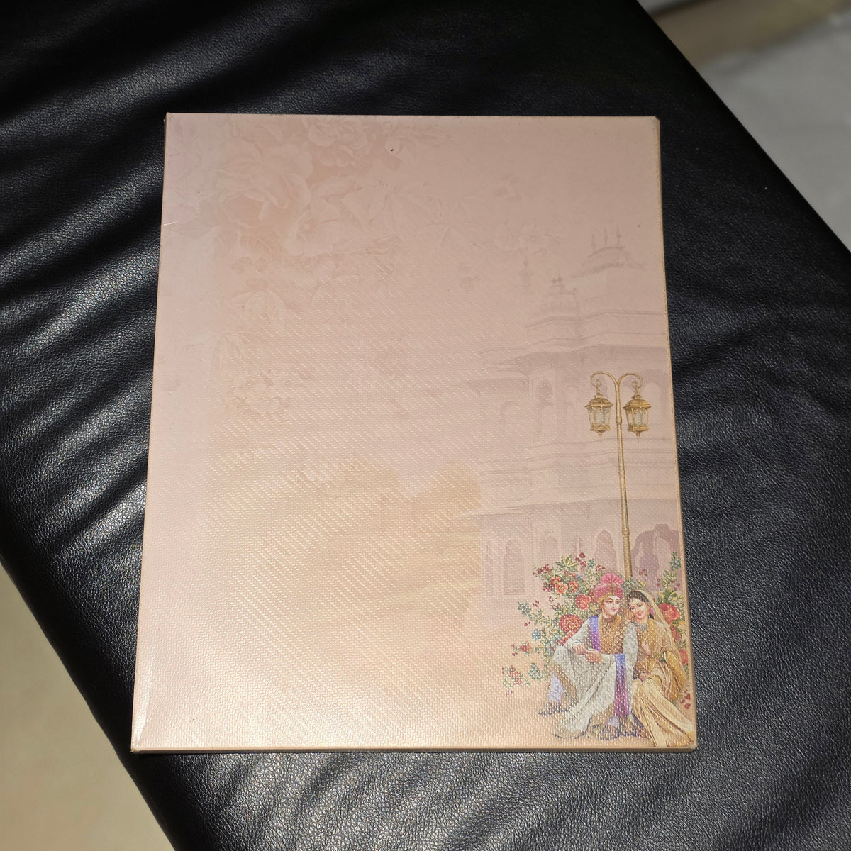 A rectangular wedding invitation card with a light peach-beige background, featuring a faint traditional pattern, a golden lamp post, and an illustration of a couple with flowers at the bottom right, resting on a black leather surface.