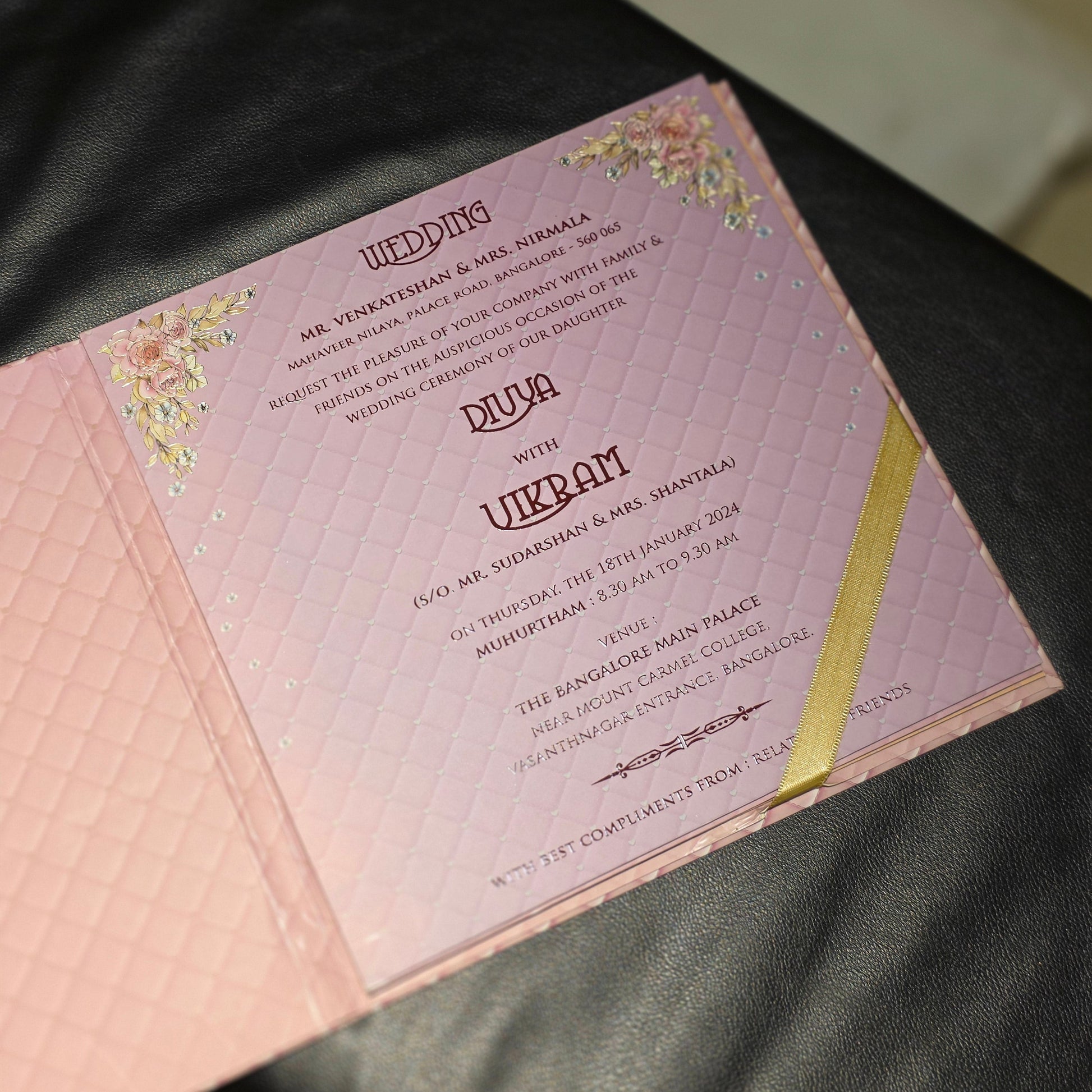 A pink wedding invitation card with gold foil floral accents and a diagonal gold ribbon, photographed on a dark textured surface.