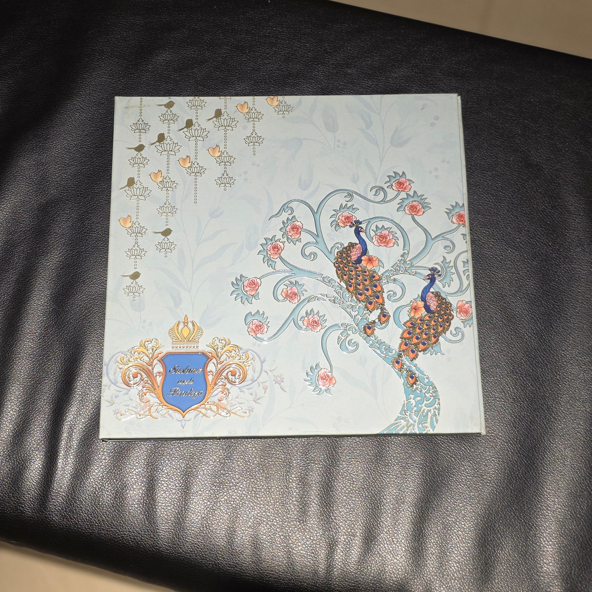 A square decorative card or print featuring peacock illustrations, floral motifs, and a blue crest with gold detailing, placed on a black leather surface.