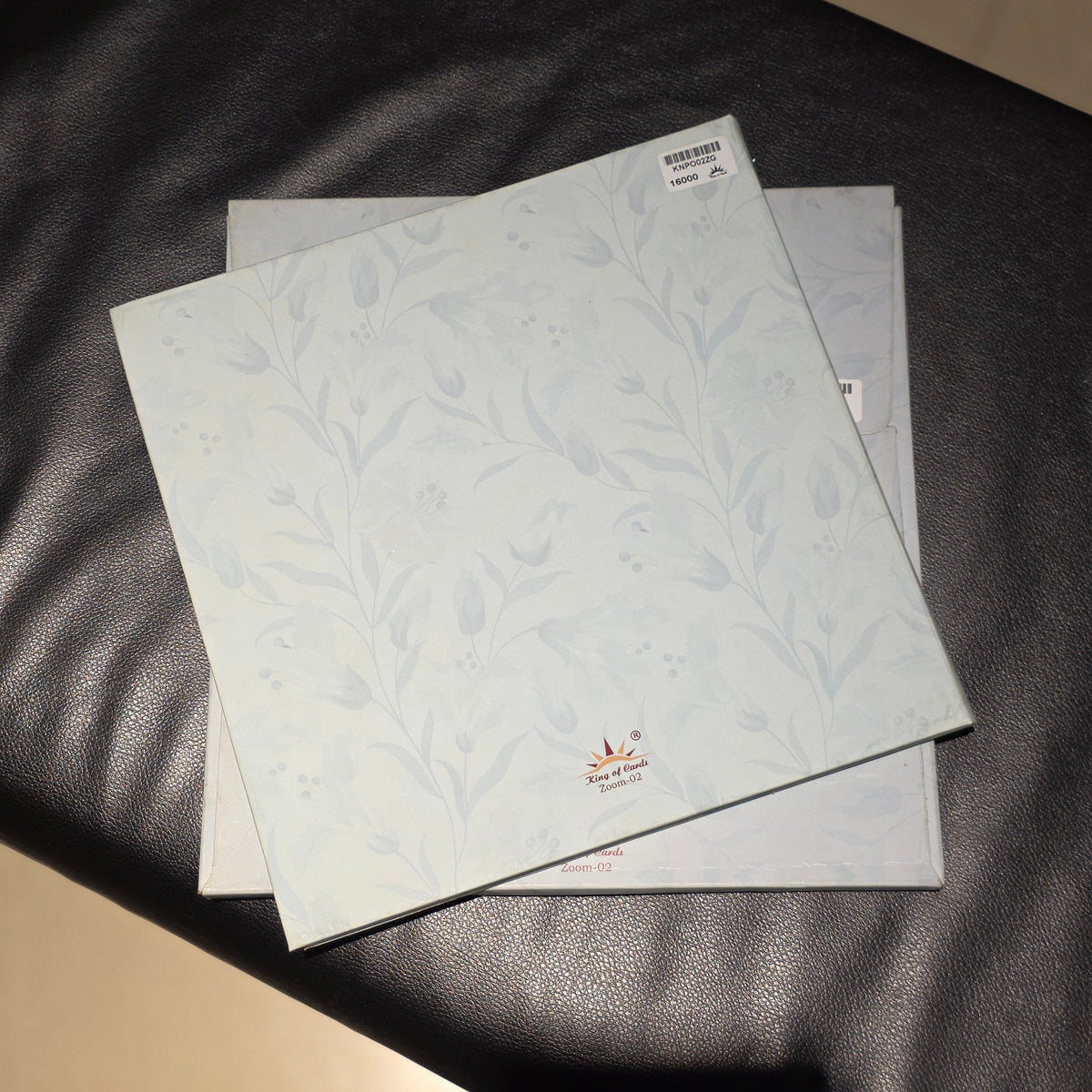 Two square decorative tiles with a pale blue and white floral pattern, stacked on a black leather surface.