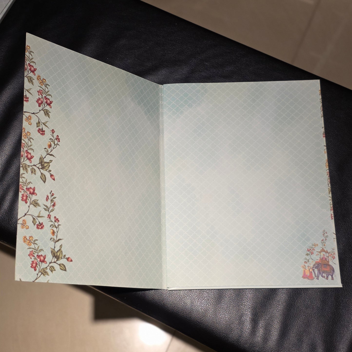 An open notebook or journal with a pale blue and white textured cover, decorated with floral patterns along the left edge and a small illustration in the bottom right corner, resting on a black leather surface.