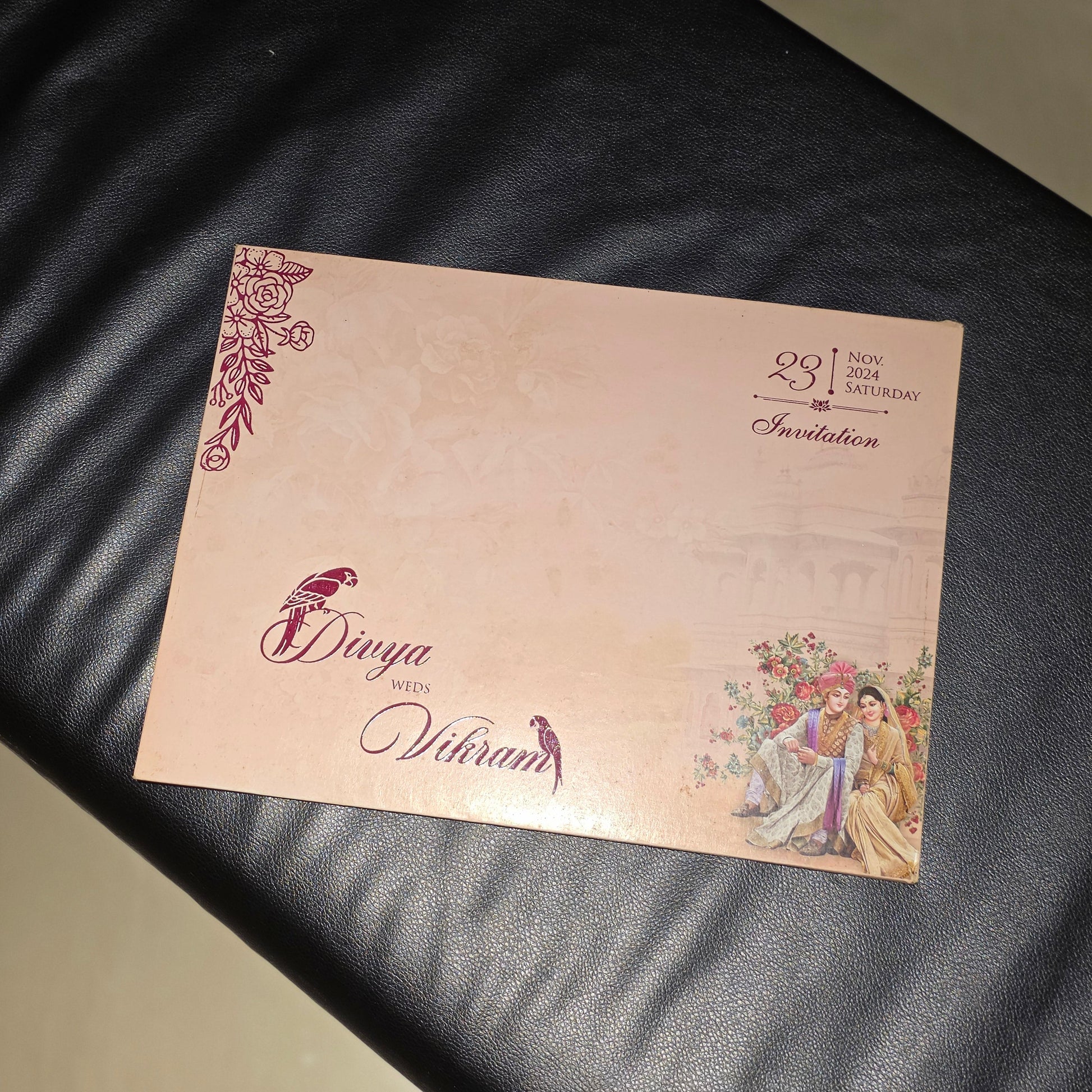 A rectangular printed invitation card with a pale pink background, decorative floral corner details, and illustrated figures in the lower right corner, placed on a black leather surface.