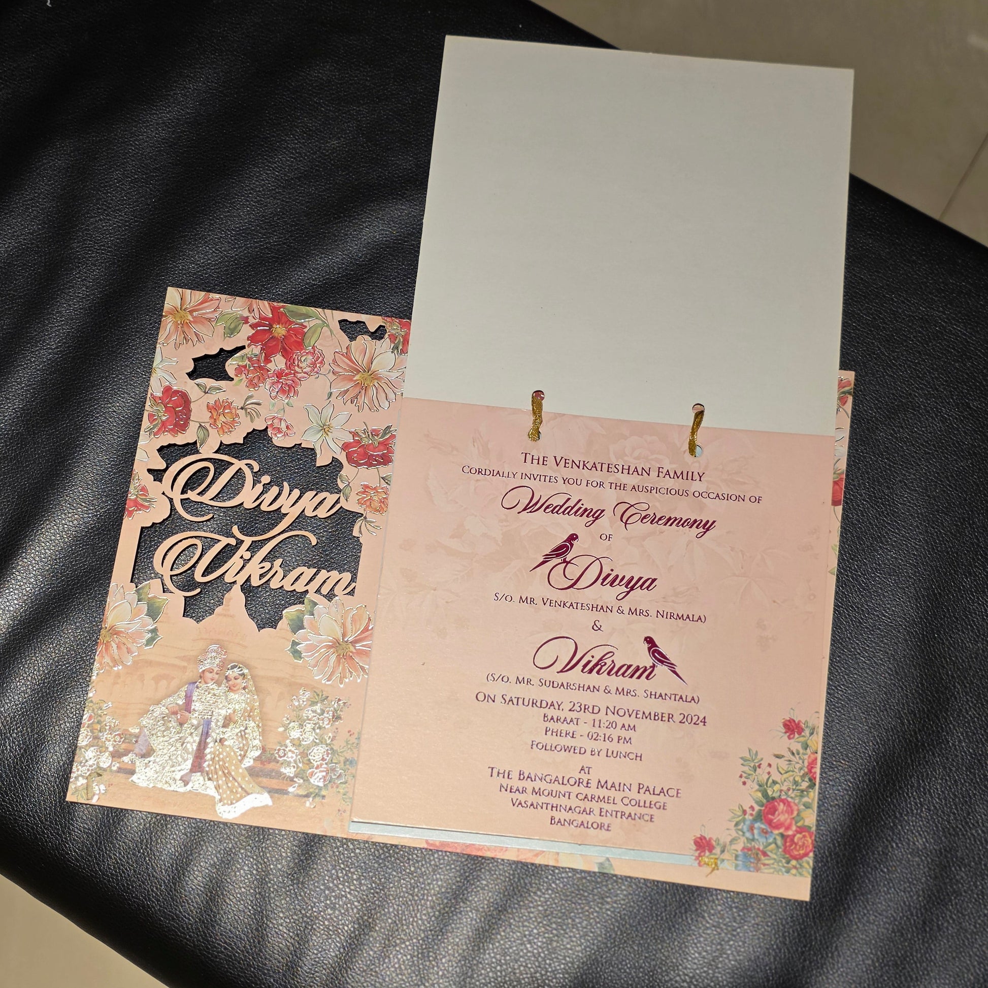 A wedding invitation suite displayed on a black leather surface, featuring a floral and metallic foil invitation card, a pink invitation booklet with gold ring binder, and a white backing card.