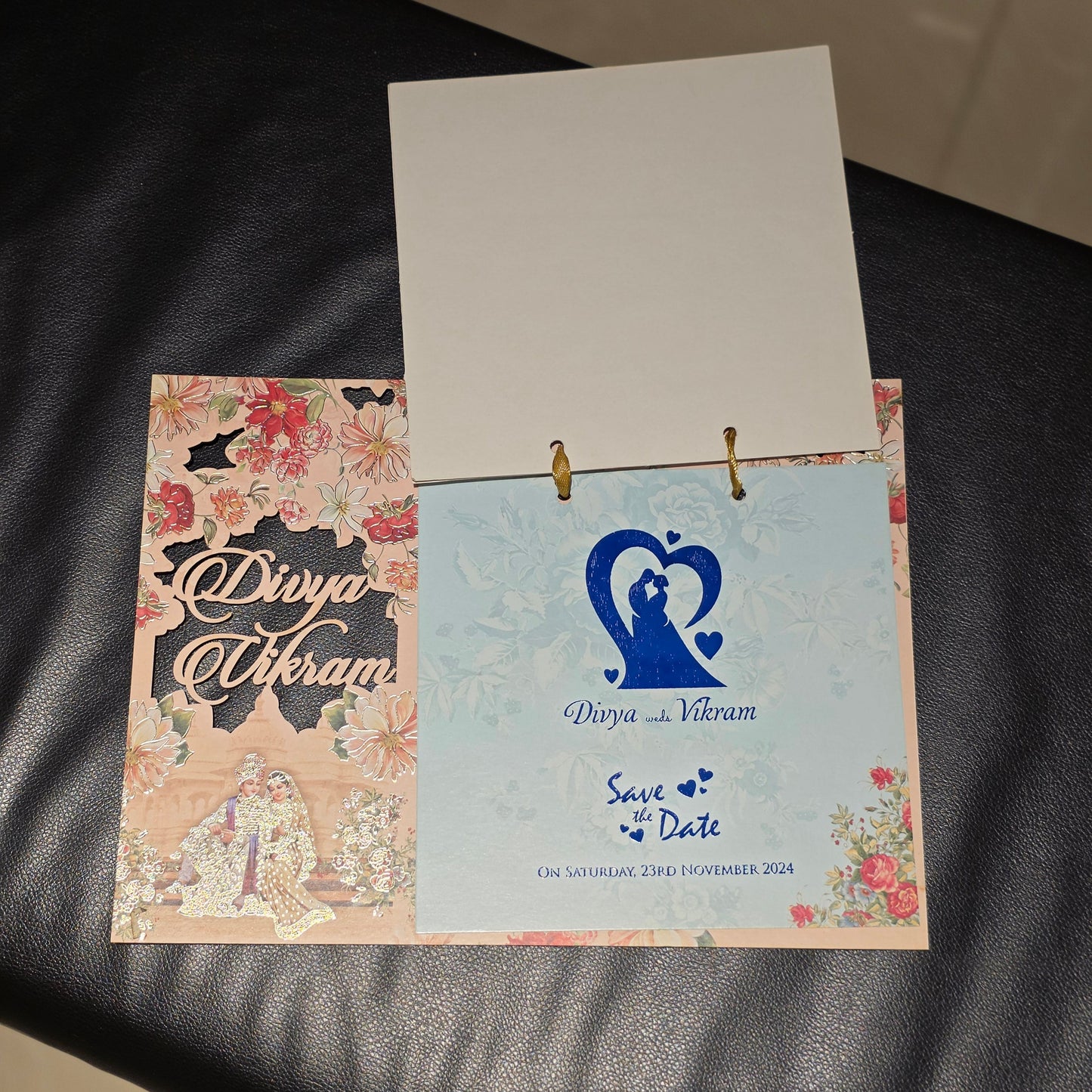 A two-page binder ring album or invitation set with a floral-patterned cover and a light blue inner page featuring a blue heart design, placed on a black leather surface.