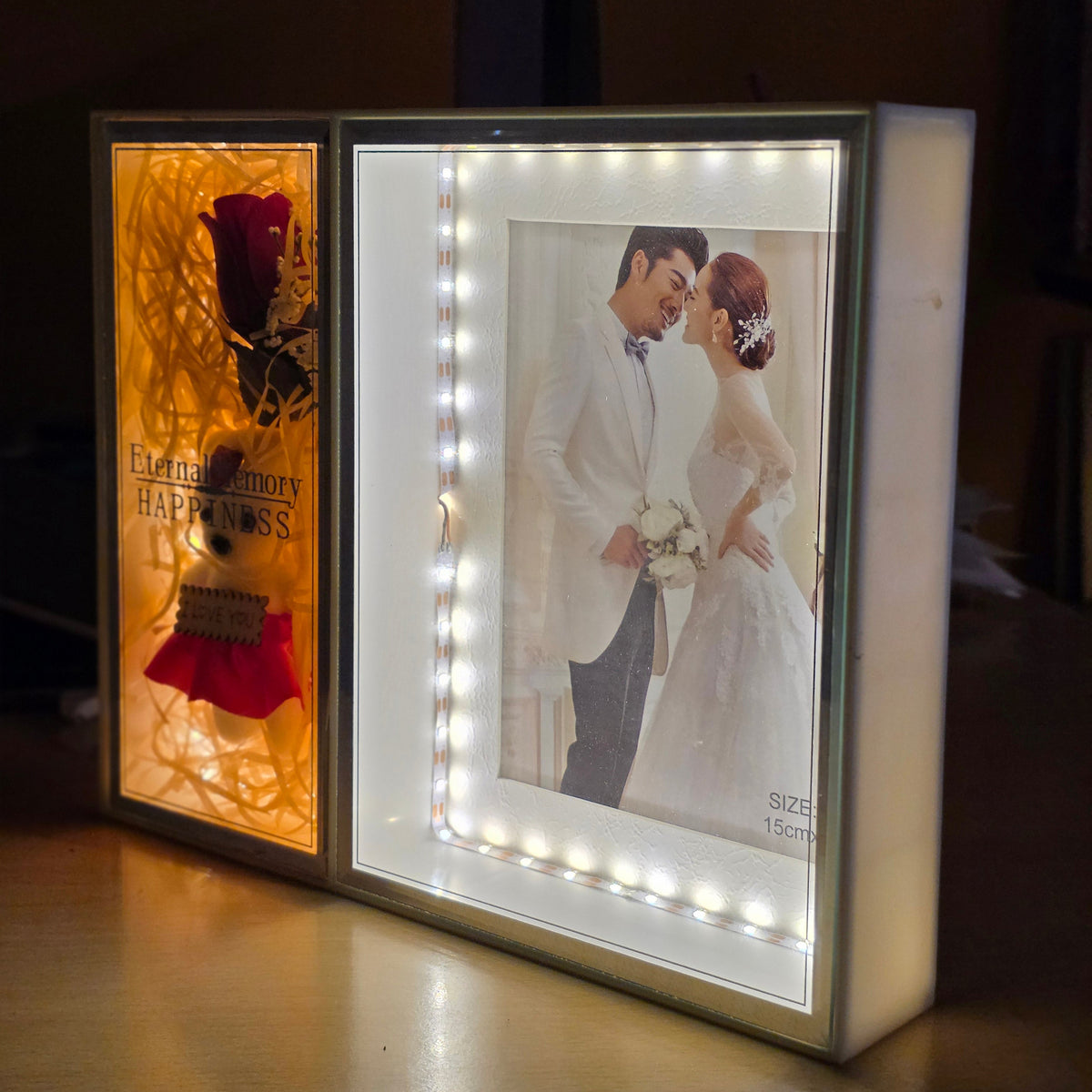 A custom LED lighted shadow box memorial display on a wooden table in a dimly lit room. The left panel features a red rose and the text 'Eternal Memory HAPPINESS', while the right panel displays a photo of a bride and groom illuminated by built-in LED lights.
