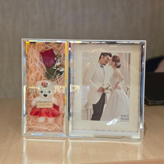 A clear acrylic double-photo frame displayed on a light wood surface against a neutral gray wall. The left photo slot contains shredded pink paper filler with a small teddy bear wearing a red dress and a red rose.