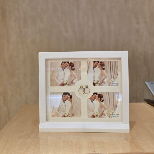 A white square photo frame with four inset wedding photos and a central ring holder, displayed on a light wood tabletop against a textured beige wall.