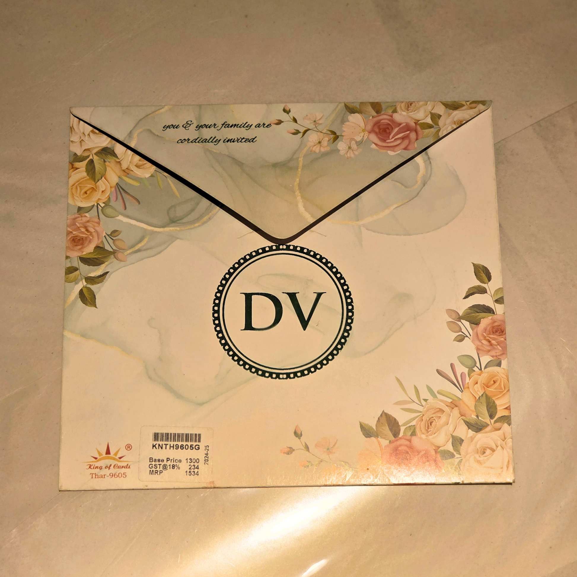 A square wedding invitation card with a floral envelope design, featuring the monogram 'DV' in the center, placed on a light beige surface.