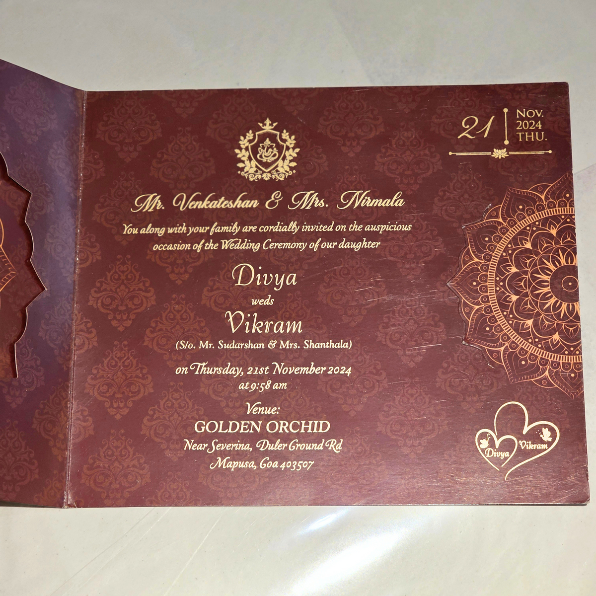 An open maroon wedding invitation card with gold foil text, ornate mandala designs, and a gold crest, photographed on a light surface.