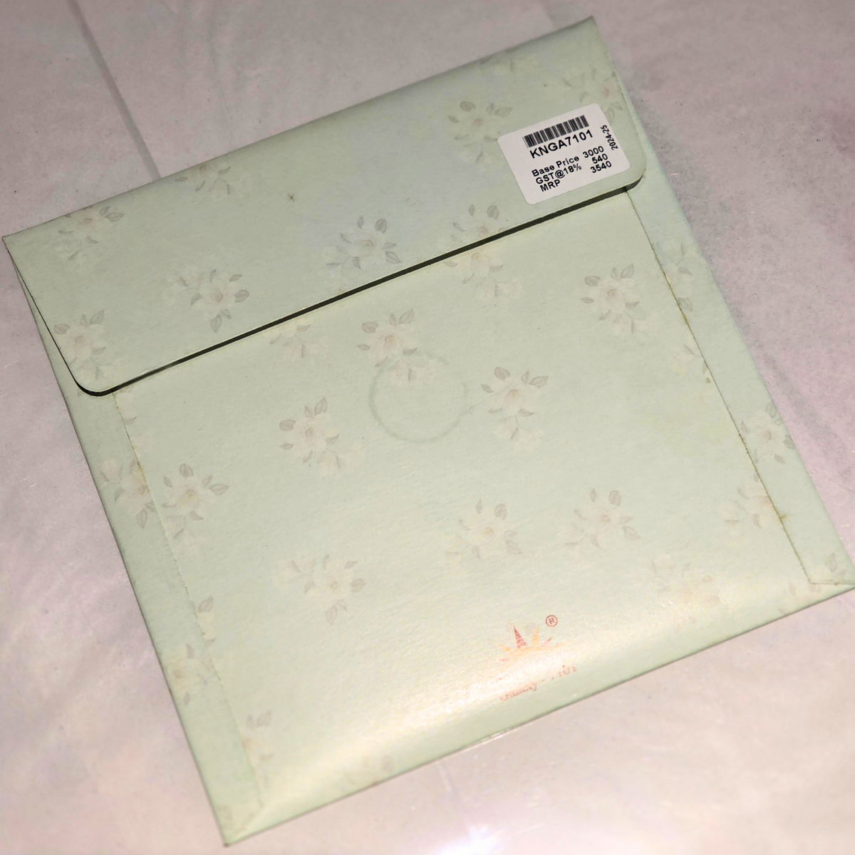 A pale mint green envelope with a subtle floral pattern, featuring a white barcode sticker on the flap, resting on a light-colored fabric surface.