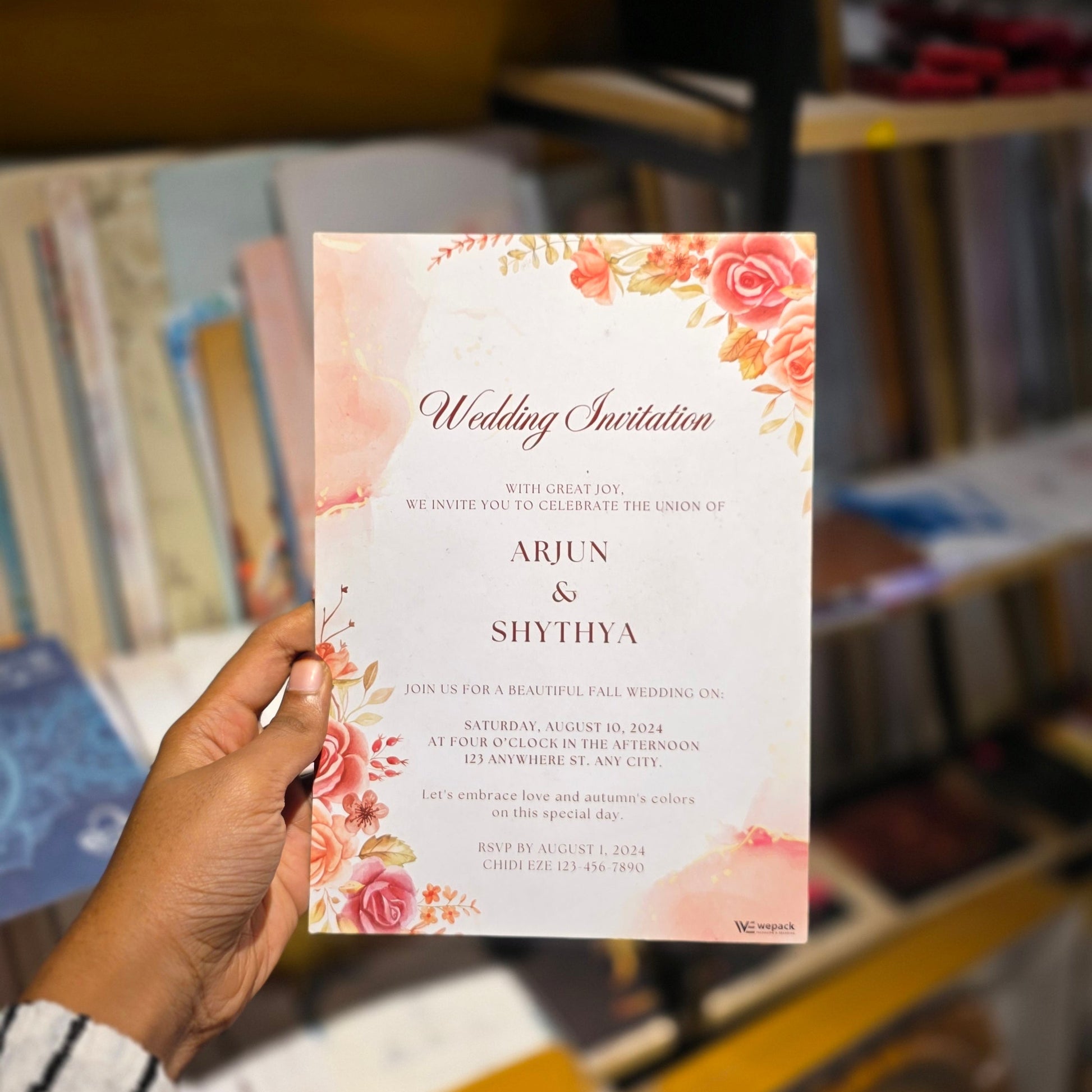 A hand holding a printed wedding invitation card with floral pink and orange illustrations on a white background, set against a blurred indoor background with shelves and objects.