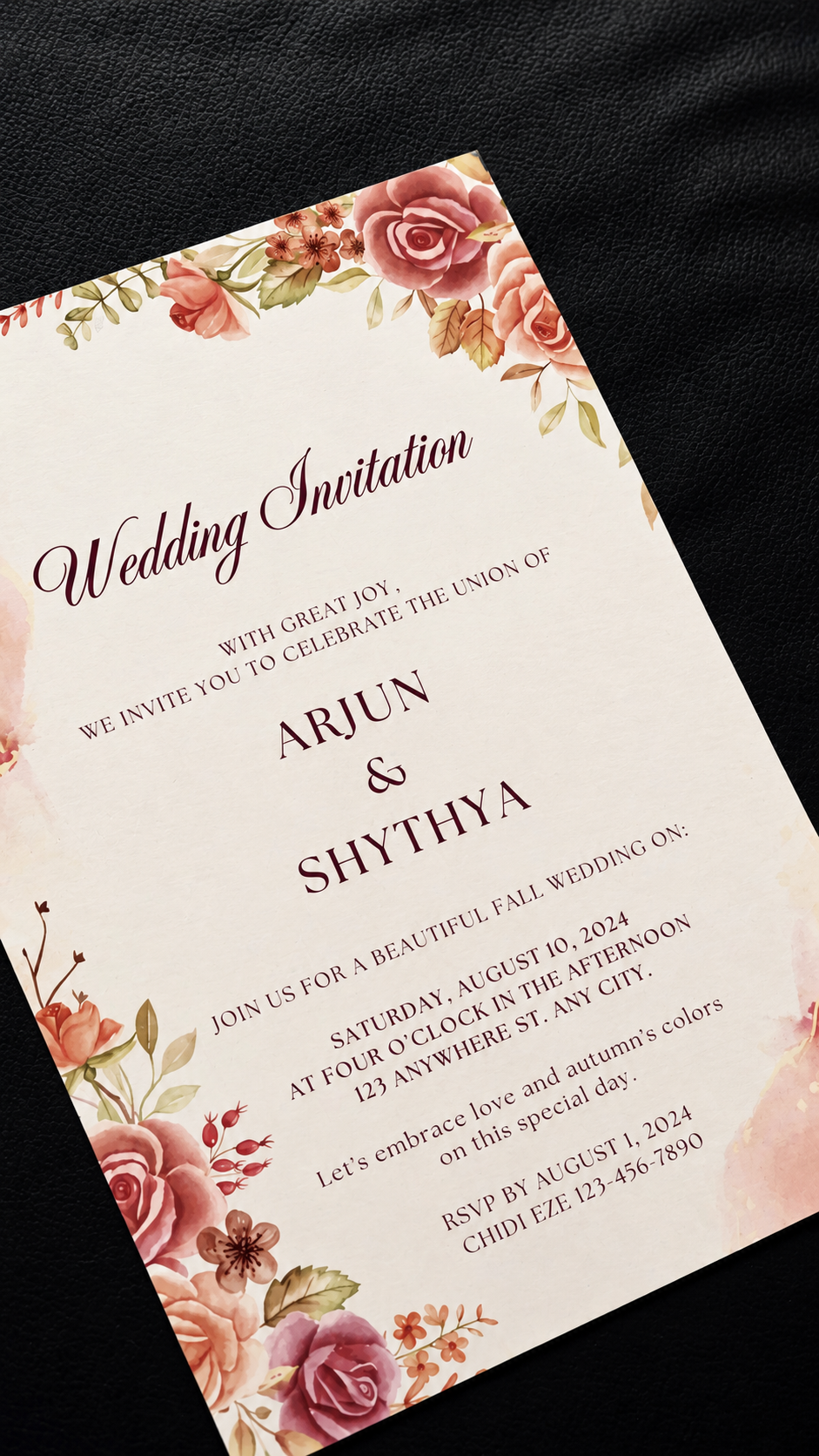 A floral wedding invitation card with pink and peach rose illustrations and elegant script text, photographed at an angle on a dark textured fabric background.