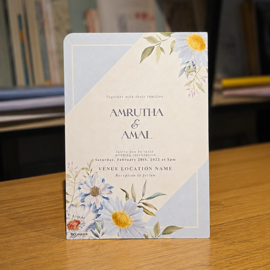 A printed invitation card with floral illustrations and the names 'AMRUTHA & AMAL' stands upright on a wooden table, with a blurred indoor background featuring shelves and books.