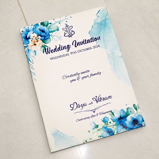 A custom wedding invitation card with blue watercolor floral designs and elegant navy text, displayed diagonally on a light beige tiled floor.
