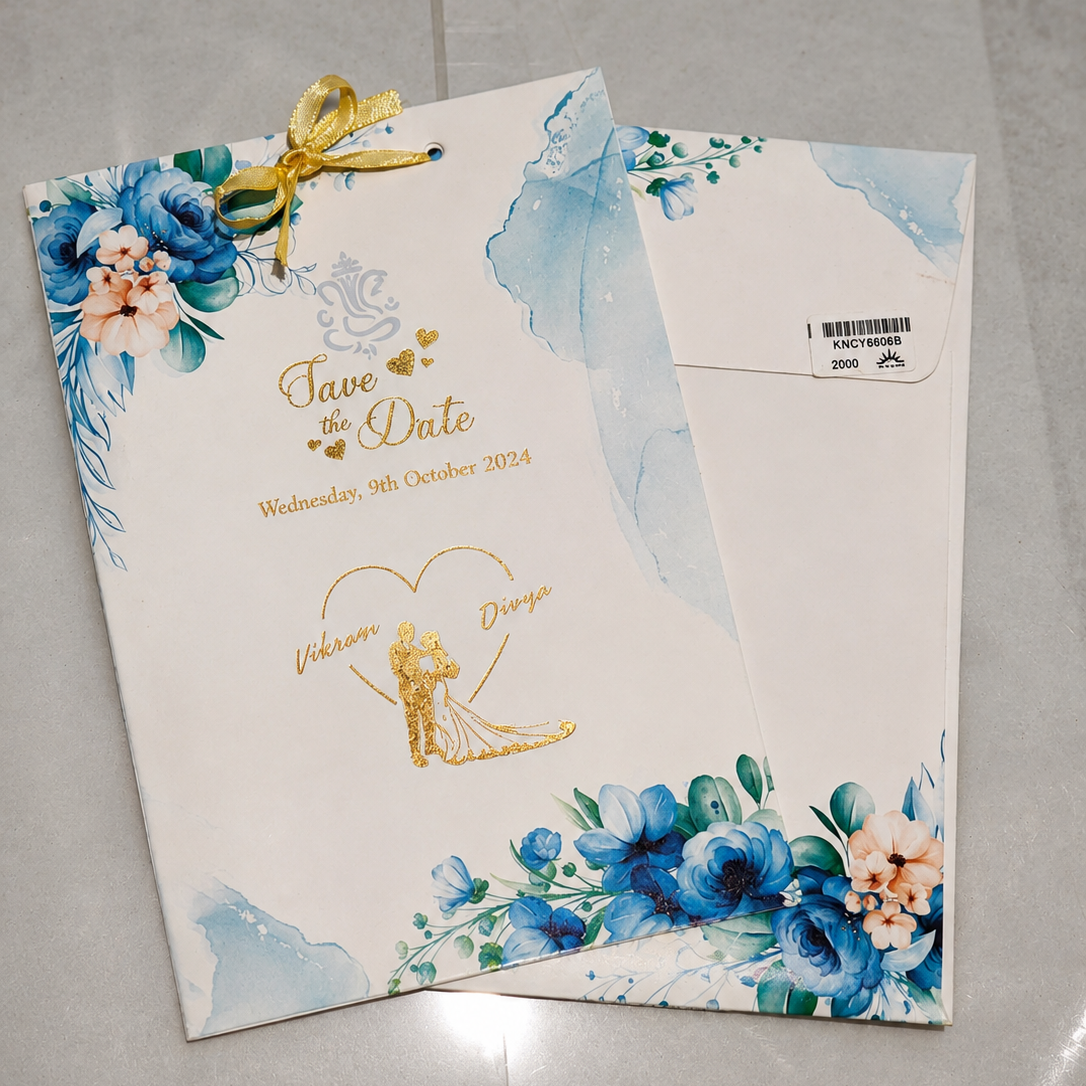 A white wedding save-the-date card and matching envelope set on a light gray tiled floor. The card features blue watercolor floral designs, gold script text reading 'Save the Date' with the date 'Wednesday, 9th October 2024', and a gold bride-and-groom illustration inside a heart.