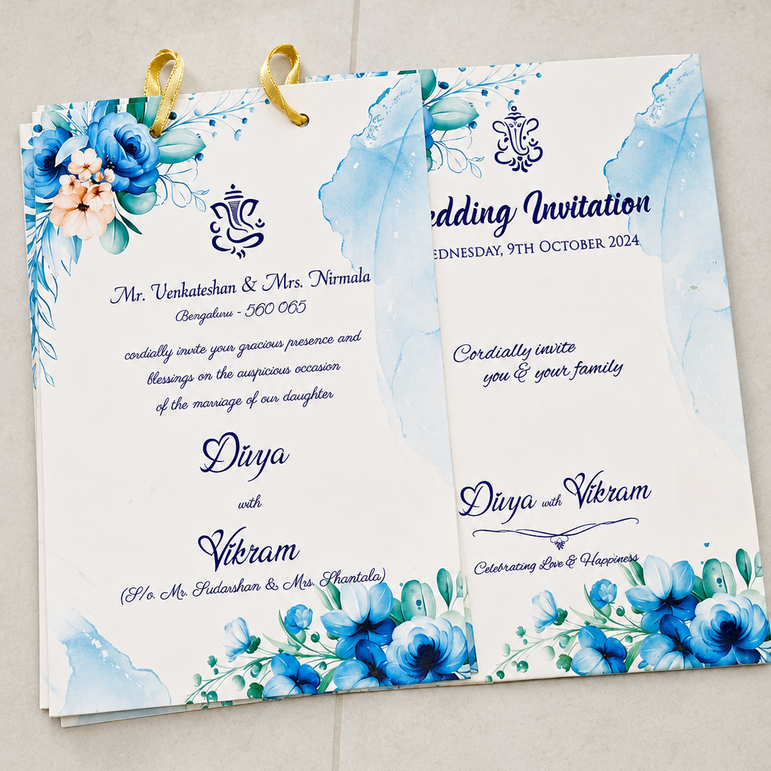 A wedding invitation card set with two overlapping white cards featuring blue watercolor floral designs, blue script text, and gold ring holders at the top, photographed on a light beige tiled surface.