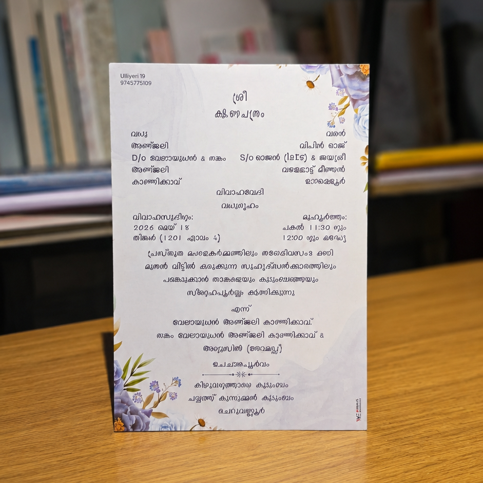 A printed card with Malayalam text and delicate floral illustrations in purple and green, placed on a wooden table with a blurred library or study background.