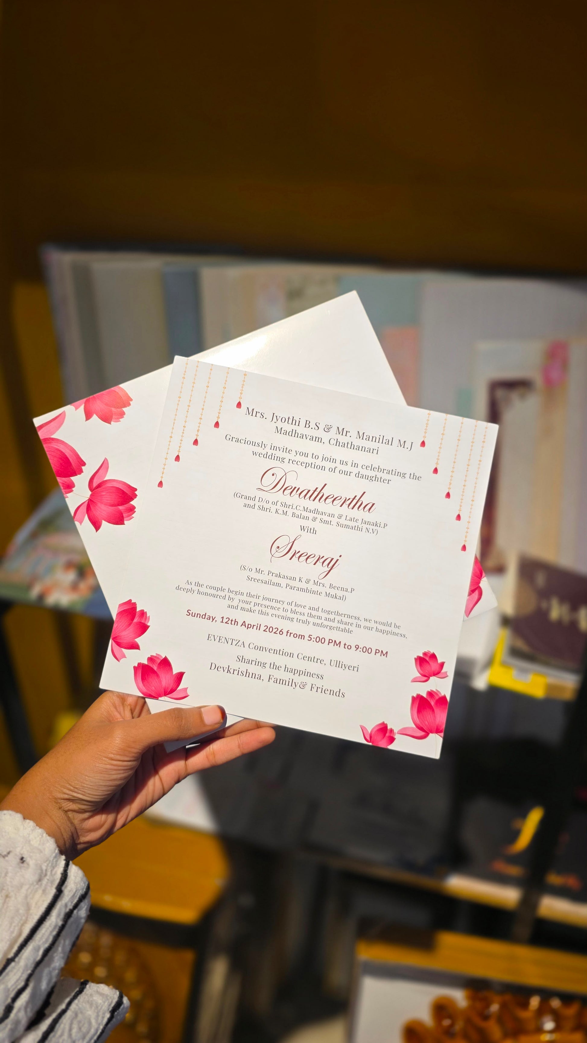 A hand holds a printed invitation card with pink floral illustrations and elegant script text, with a matching envelope behind it. The background is a softly blurred indoor setting.