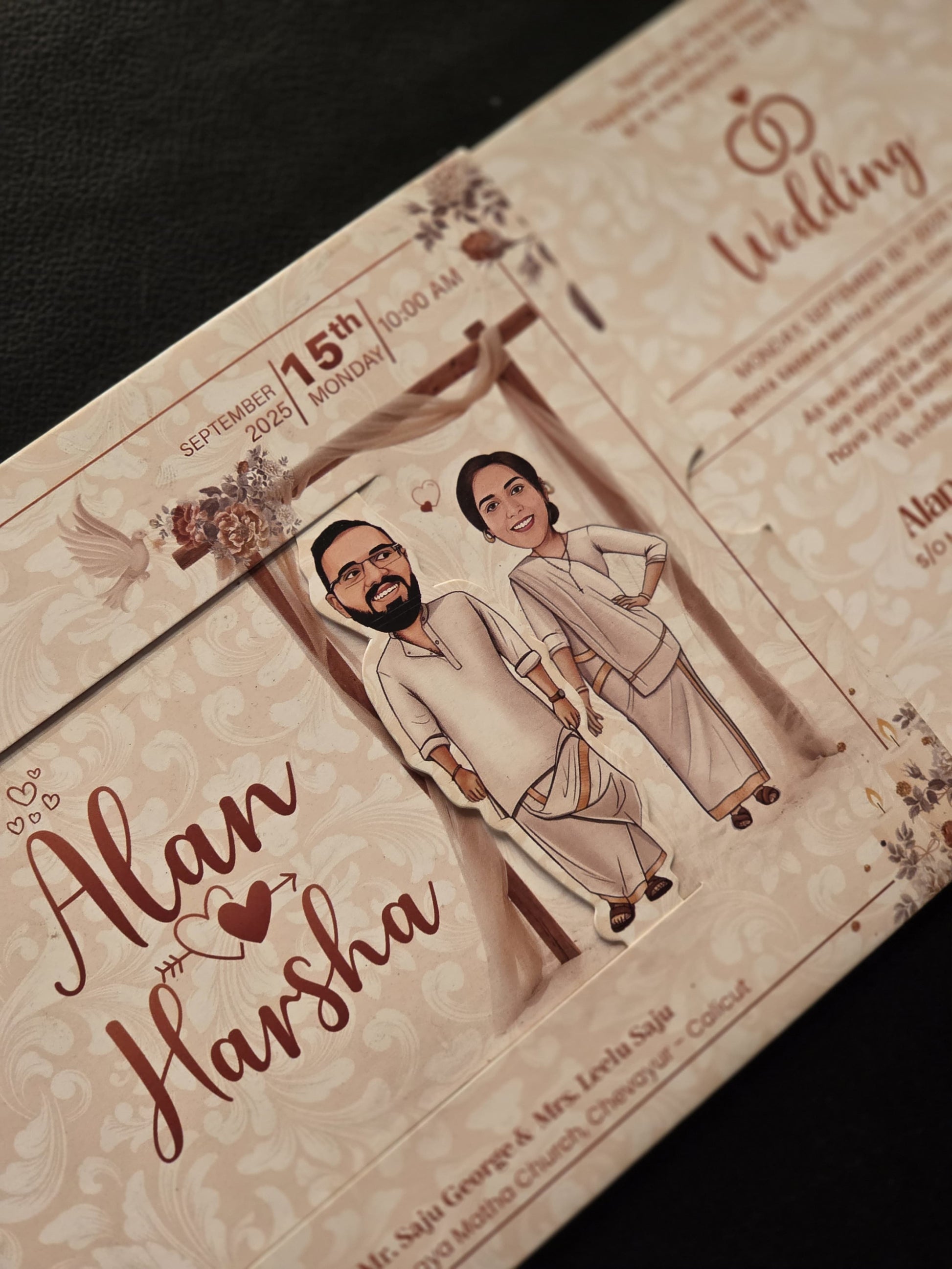 A close-up of a wedding invitation card featuring a cartoon-style illustration of a couple in traditional clothing, with the names 'Alan' and 'Harsha' in elegant cursive script on a cream-colored patterned background.