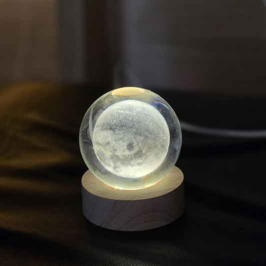 A glowing moon sphere lamp made of clear acrylic, containing a detailed moon print, resting on a round light wood base against a dark, softly blurred background.